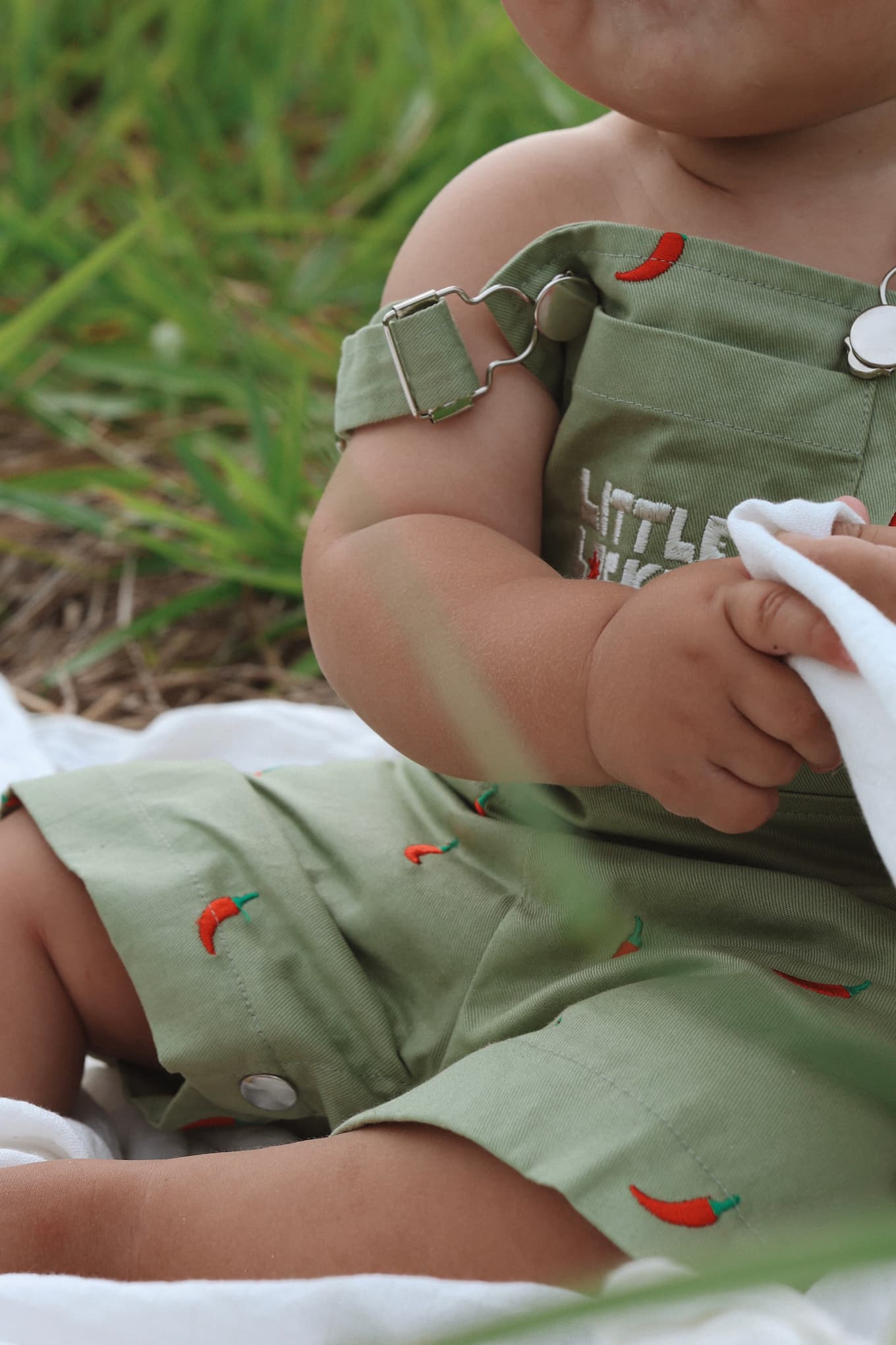 Close up of a baby wearing sage green overalls with red chilli pattern and 'Little Locker' logo. Ethically made from 100% cotton.