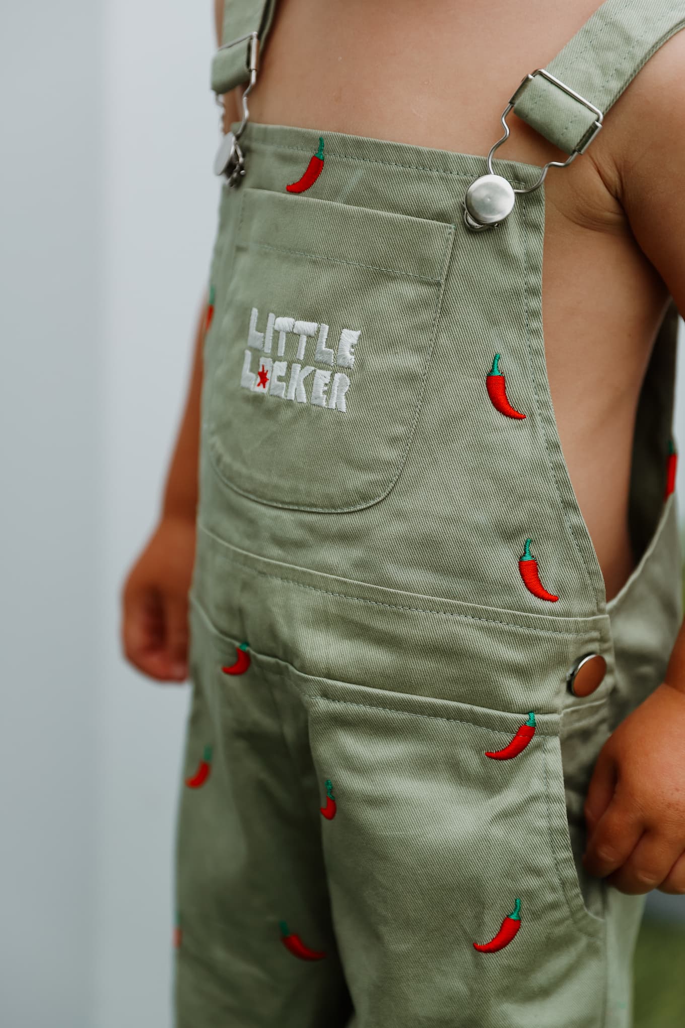 Close up of saeg green cotton overalls with 'Little Locker' logo and chili pepper designs.
