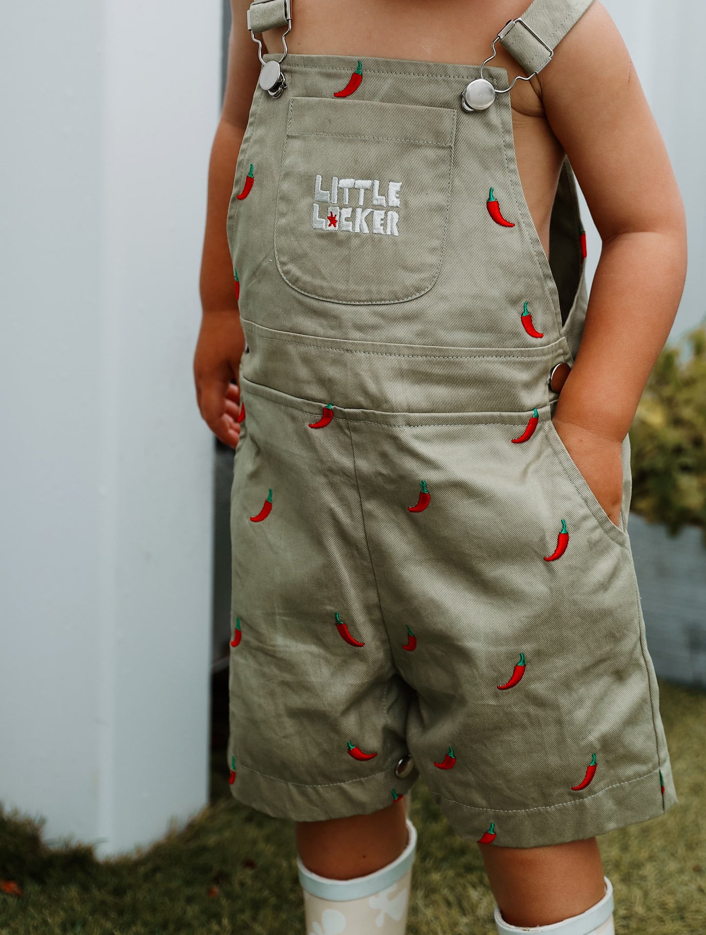 Toddler child wearing sage green overalls with red chili pepper pattern and 'Little Locker' logo.