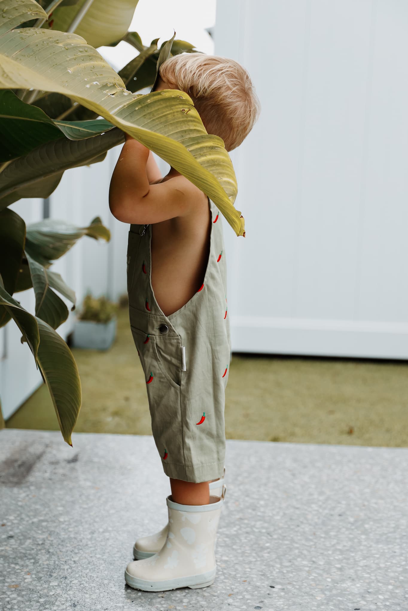 Toddler boy wearing sage green overalls with red chili pepper pattern, ethically made from 100% cotton.
