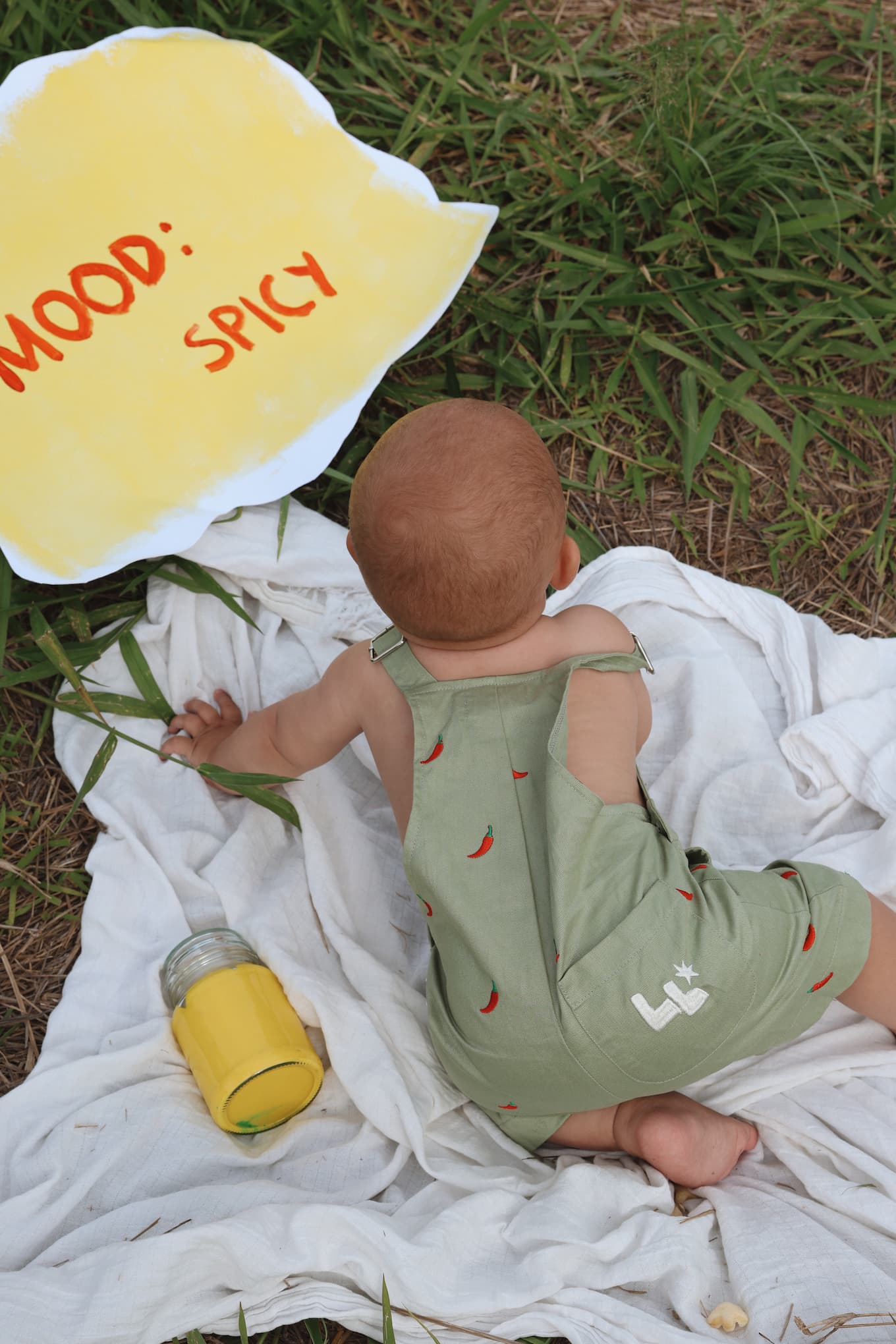 Baby boy wearing sage green overalls with red chilli pattern and 'Little Locker' logo.