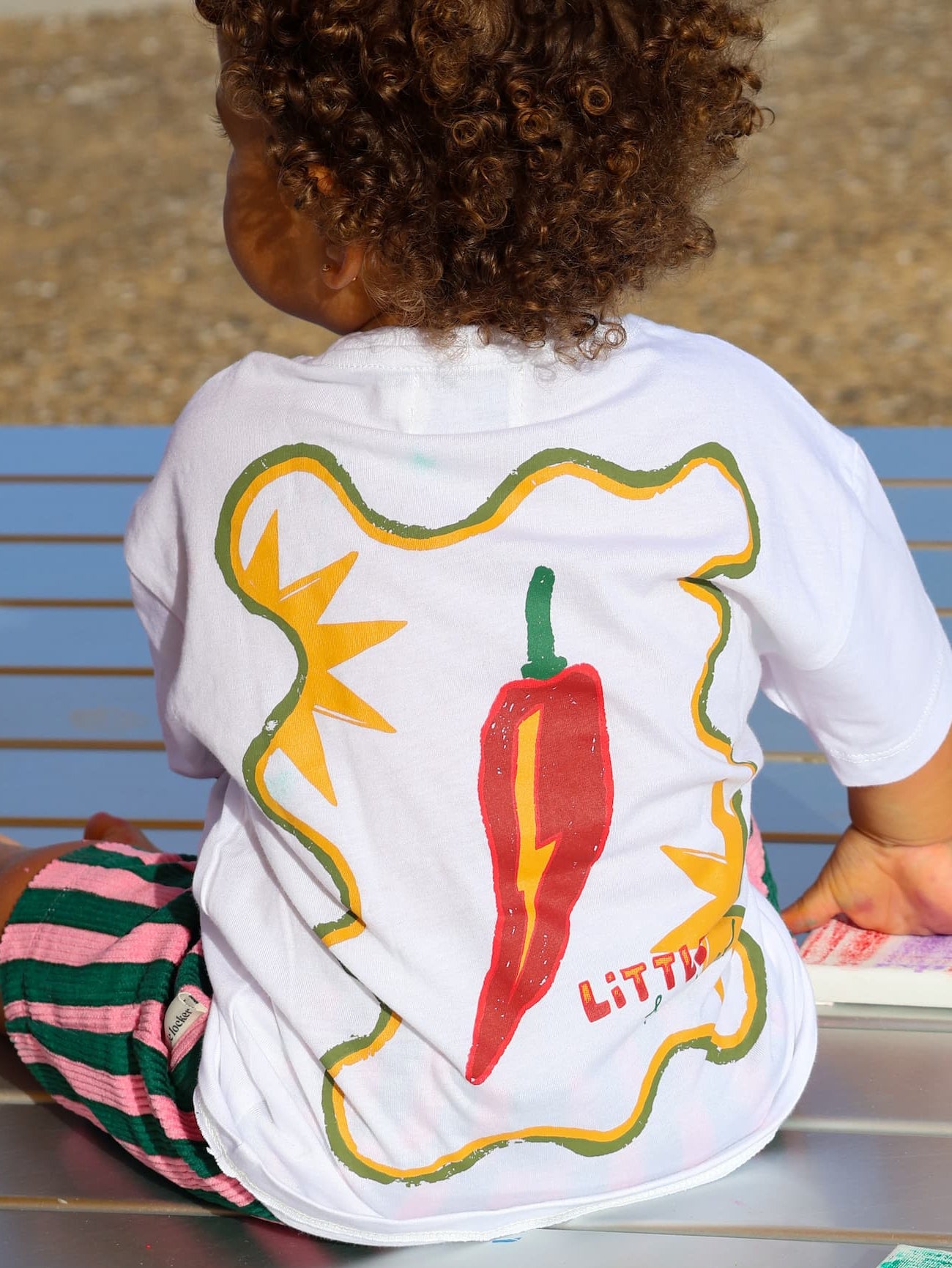 Child wearing Little Locker white t-shirt with colourful chilli design on back with striped shorts.
