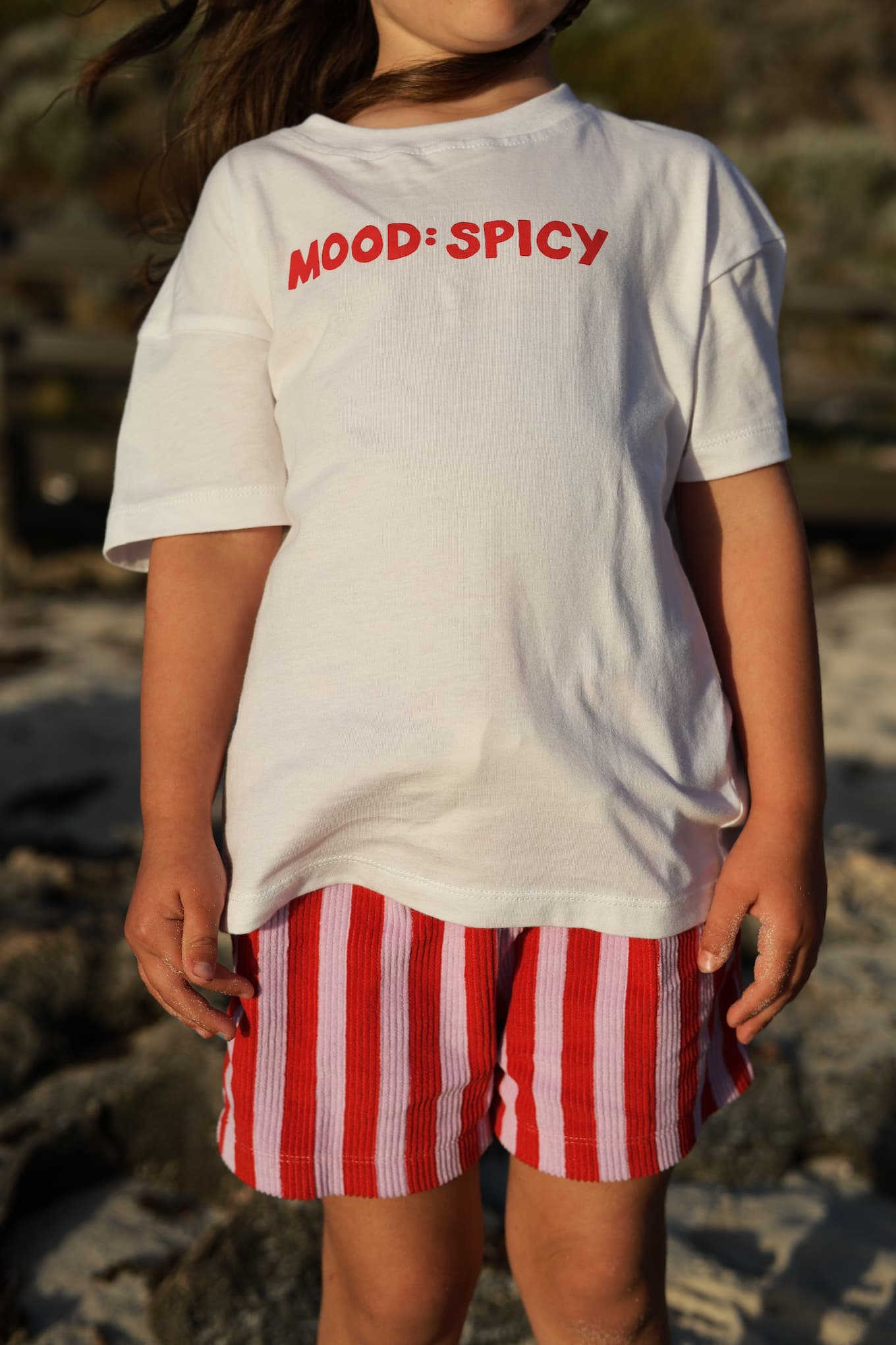 Child wearing a Little Locker 100% cotton t-shirt with 'Mood: Spicy' text and red and purple striped shorts.