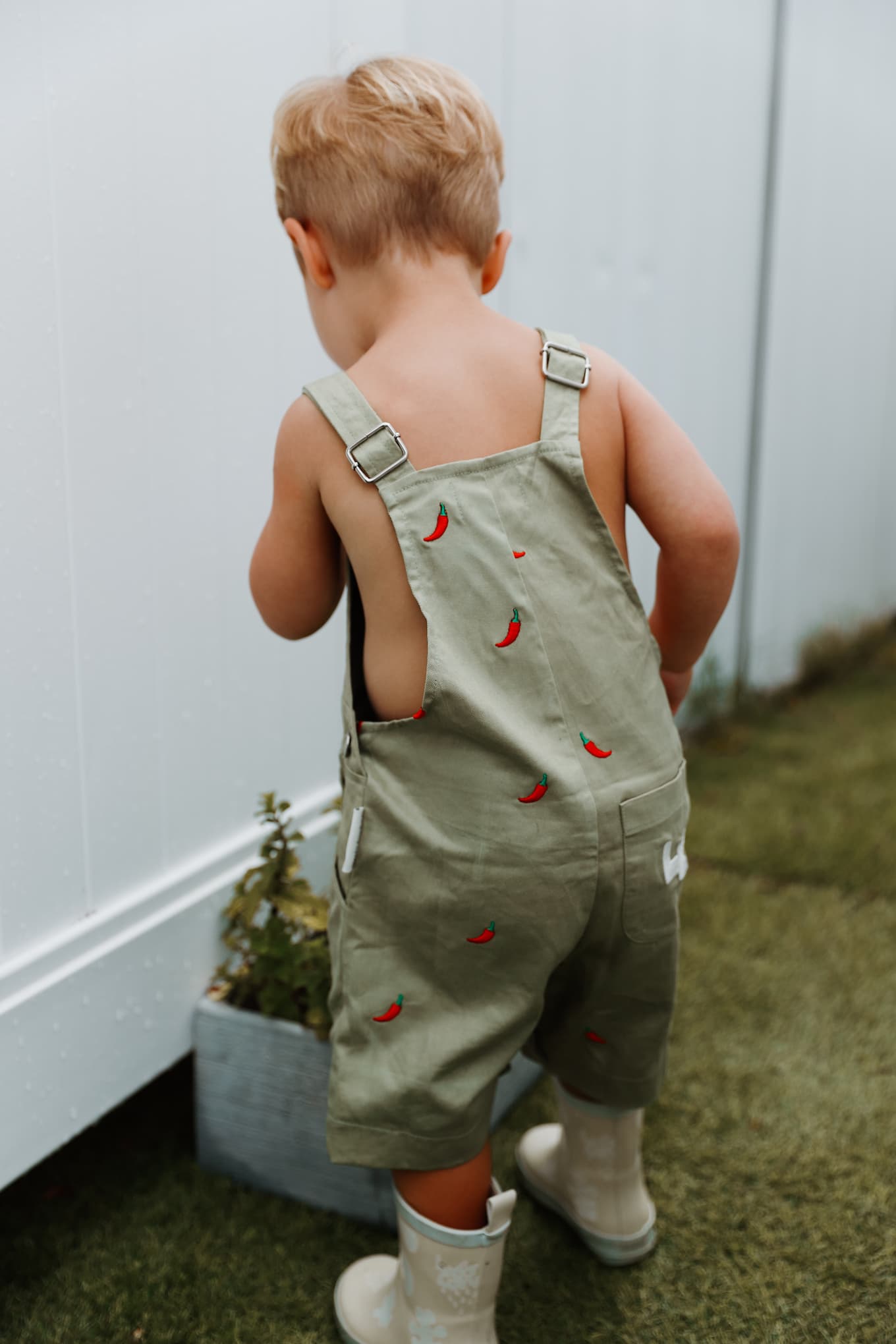 Little boy wearing sage green cotton overalls with red chillies embroidered throughout.