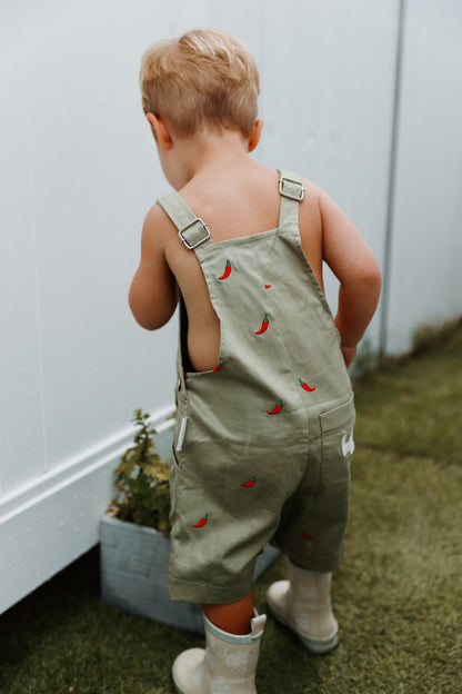 Little boy wearing sage green cotton overalls with red chillies embroidered throughout.